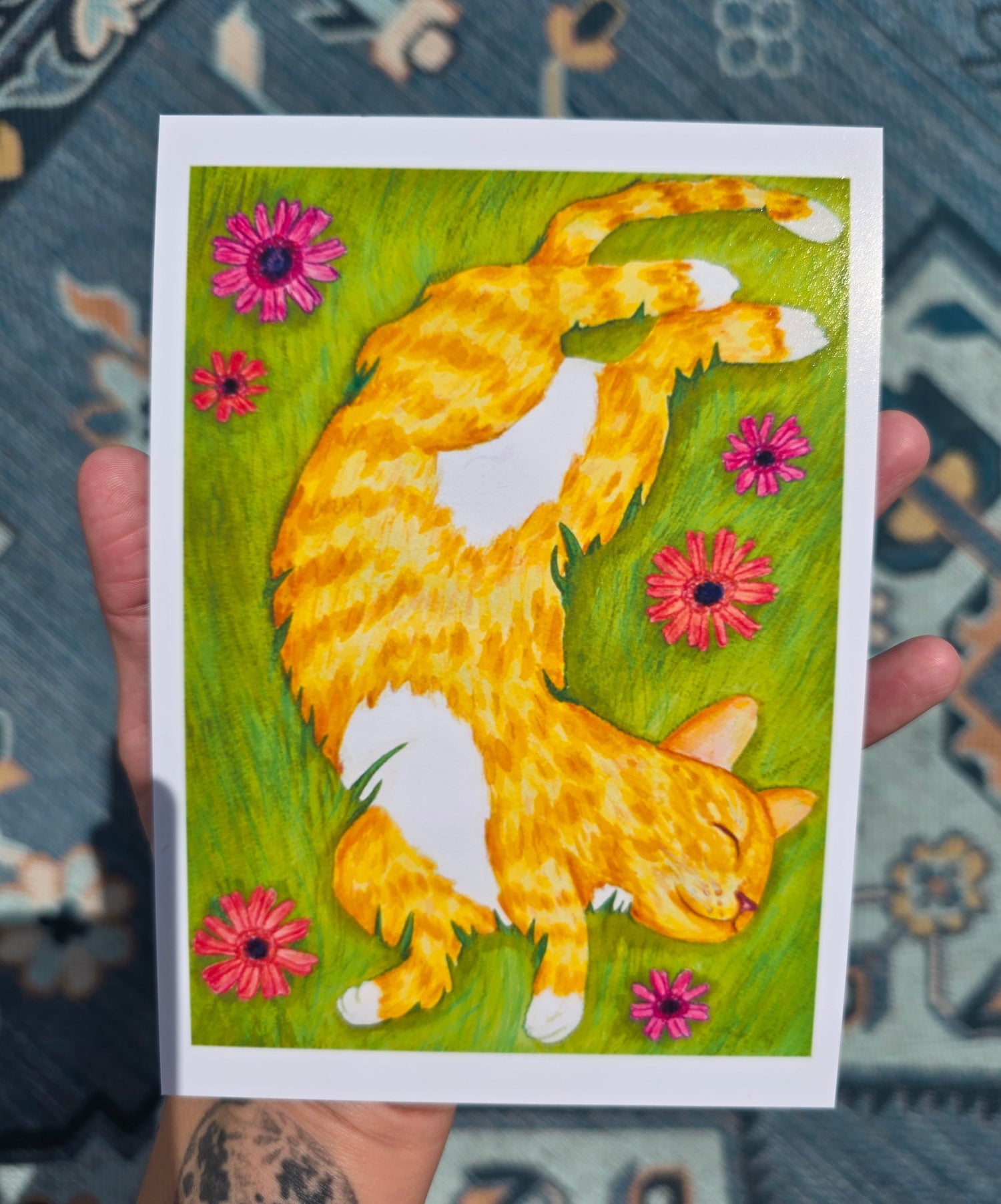 An art print being held by the artist shows an orange cat sleeping on its back in a grassy, flowery field. There are six flowers surrounding the sleeping cat. The cat looks content and comfy.